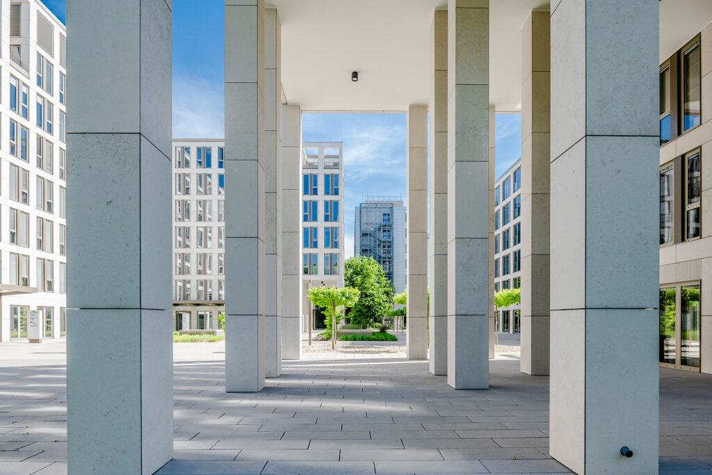 Innenhof des BMH-Gebäudekomplexes: Blick durch eine Reihe heller Steinsäulen auf einen begrünten Innenhof mit Bürogebäuden und Bäumen unter blauem Himmel.