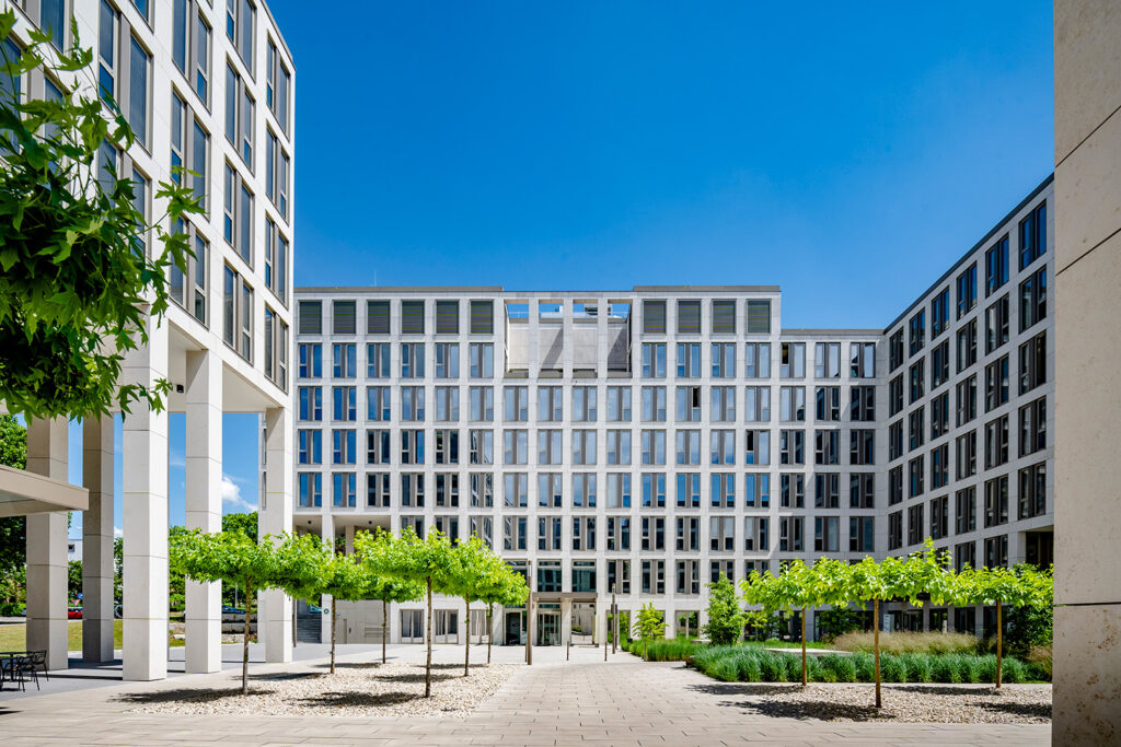 Innenhof des mehrgeschossigen BMH-Bürokomplexes mit heller Fassade, im Vordergrund Platanen und eine begrünte Fläche.
