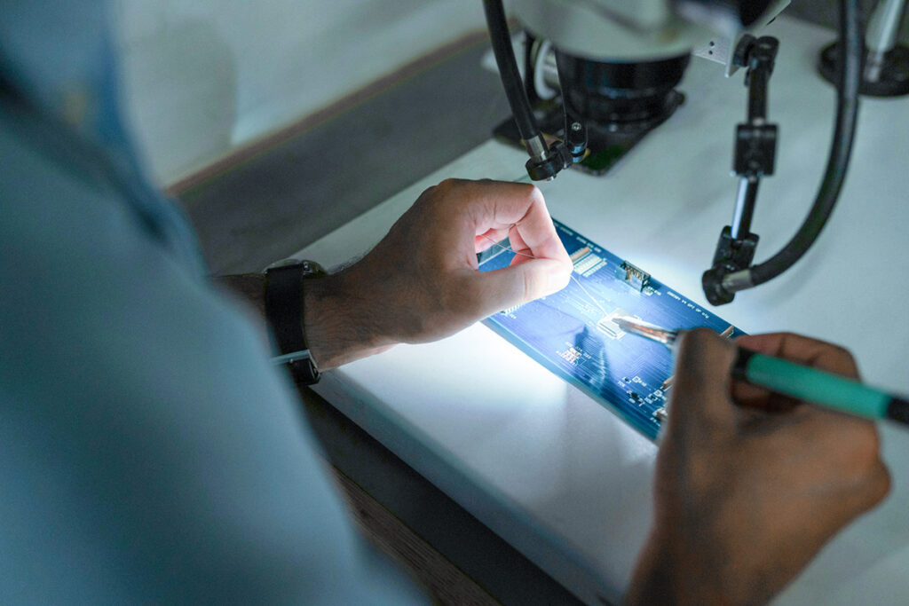 Nahaufnahme einer Person, die mit Werkzeugen an einer elektronischen Platine unter einem Mikroskop arbeitet.